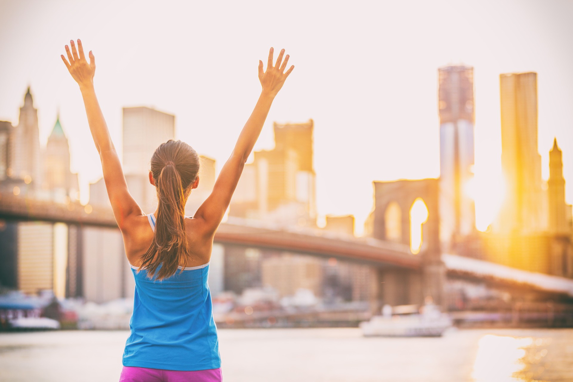 Happy cheering fit woman achieving fitness goal in New York City with arms up at sunset and Brooklyn Bridge. Girl runner joyful and excited after running.